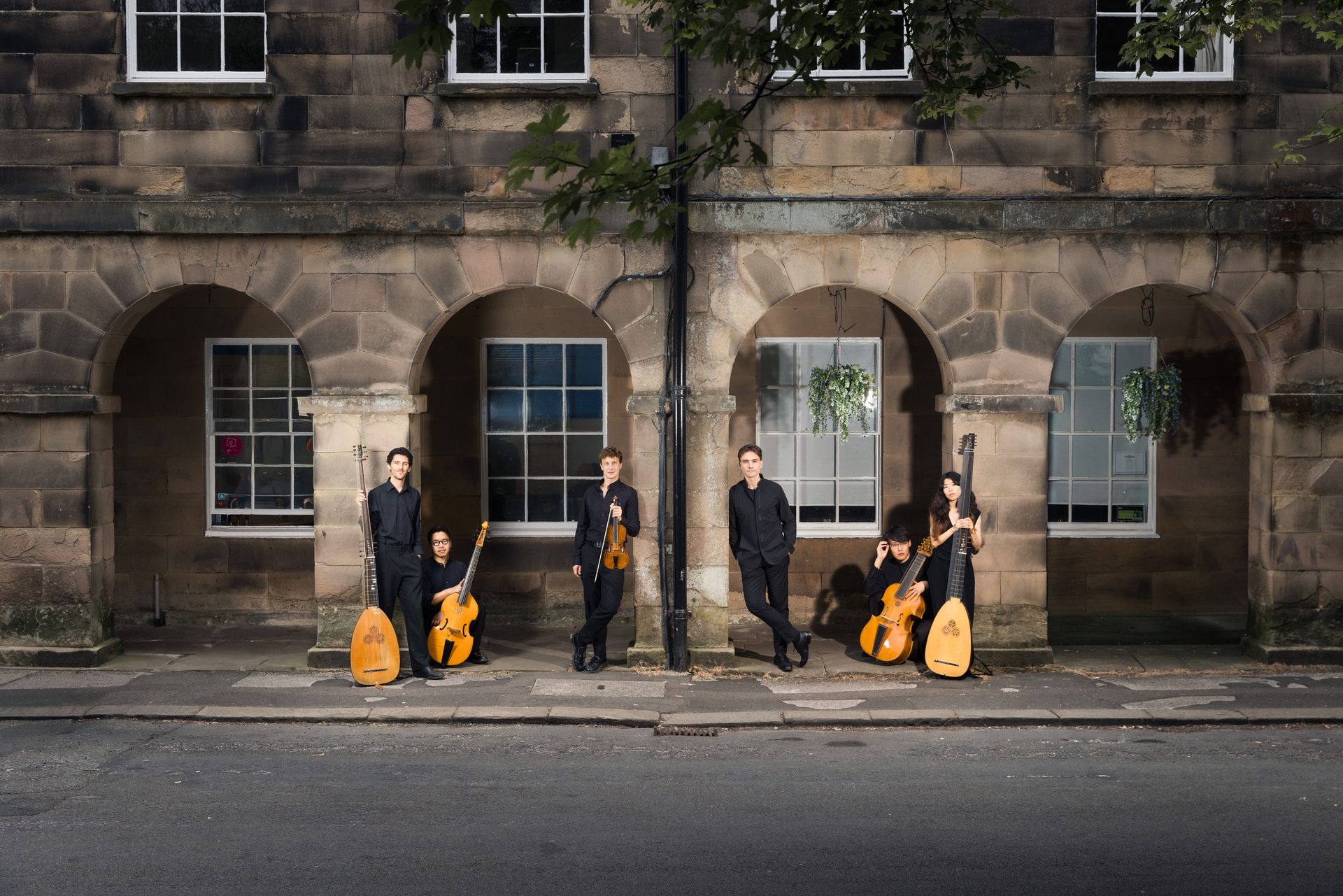 The musicians of Música d'Outrora, dressed in black with violin, viols, theorbo and archlute, standing beneath stone arches at dusk